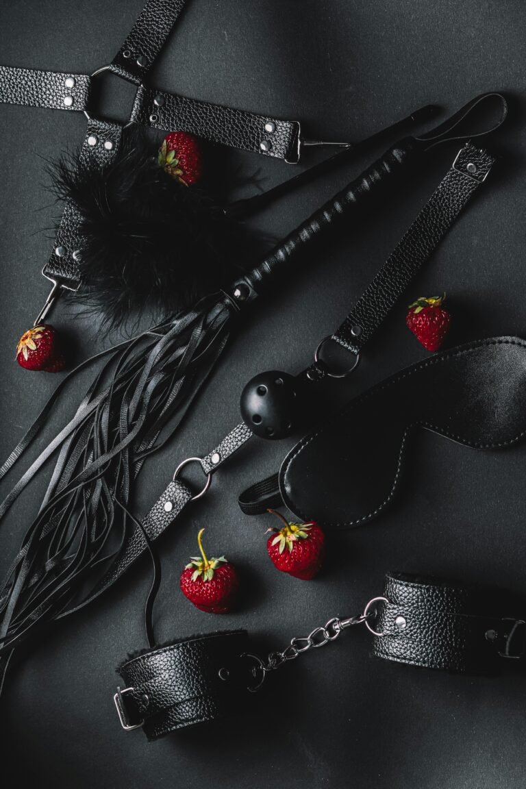 A collection of BDSM accessories, including a flogger, leather cuffs, blindfold, ball gag, and harness, arranged on a dark surface with scattered strawberries.